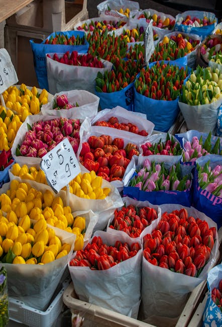 An outdoor flower market stall displaying numerous large baskets filled with vibrant tulips in various colors, including yellow, pink, red, orange, and purple. The tulips are arranged in tightly packe