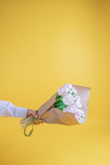 A person in a white shirt is holding a bouquet of fresh, light pink tulips wrapped in light brown paper with a green ribbon, presented against a vibrant yellow background. The tulips have smooth, deli