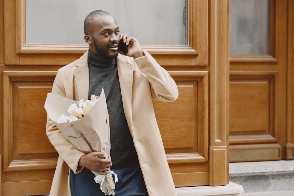 A man with dark skin, a trimmed beard, and a smile is standing outdoors in front of a wooden-paneled building, holding a bouquet of white and cream-colored flowers wrapped in brown paper. He is wearin