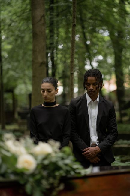 The image depicts two individuals, a woman and a man, standing in a peaceful outdoor setting with lush green trees and soft natural light. The woman, with dark hair pulled back, wears a black long-sle