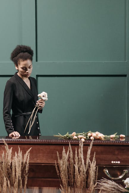 A young woman dressed in black, with curly hair, stands beside a wooden casket holding a small white floral tribute. In front of her, on the surface of the casket, there are several pale pink roses an