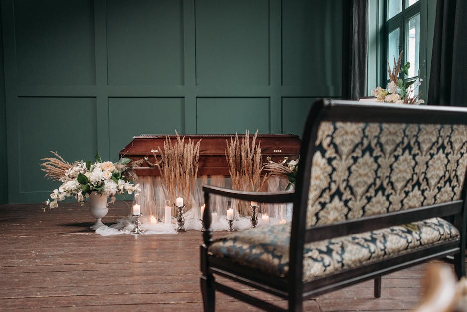 A funeral floral arrangement set on a polished wooden surface against a dark green paneled wall, featuring a large white ceramic vase filled with white roses, lilies, and greenery, complemented by tal