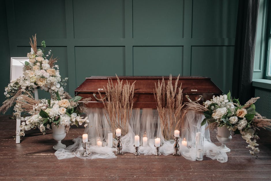 A funeral floral arrangement displayed on a wooden floor against a dark green paneled wall, consisting of two large white vases filled with cream and white roses, hydrangeas, and other light-colored f