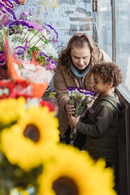 A woman and a young girl are standing by a large window in a florist shop, surrounded by vibrant flower arrangements. The woman, with light brown curly hair and a light blue sweater, is smiling as she