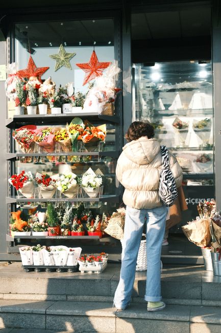 A woman stands outside a flower shop, looking at a display of vibrant floral arrangements and holiday decorations. The display features a variety of fresh flowers, including red, white, pink, and yell