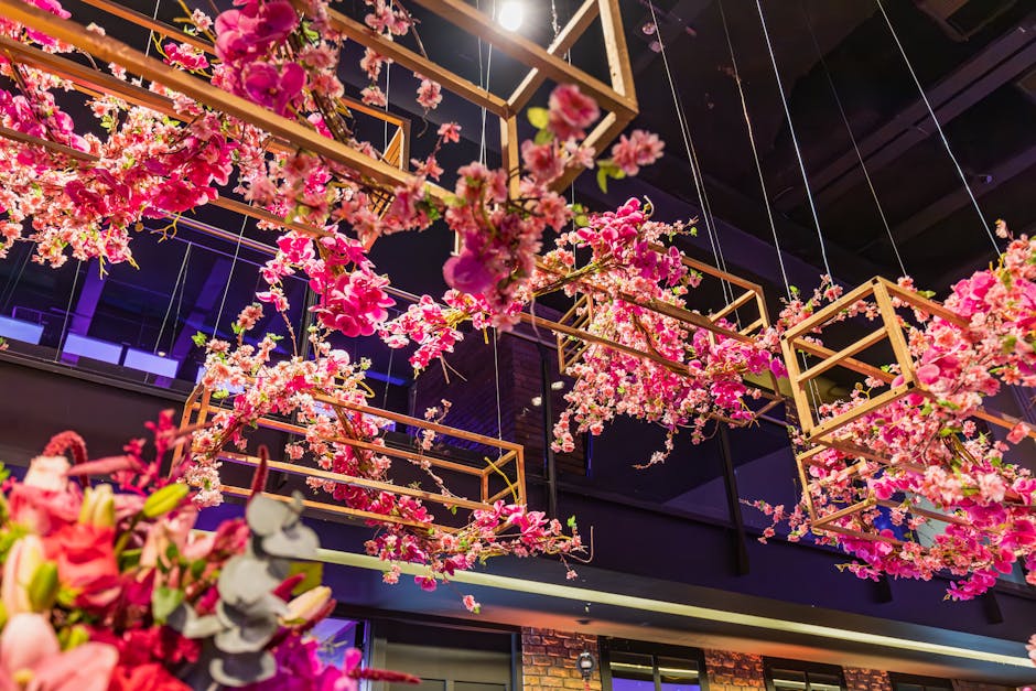 An elaborate floral installation featuring pink cherry blossom branches and clusters of light pink and dark pink flowers suspended within wooden geometric frames hanging from the ceiling. The arrangem