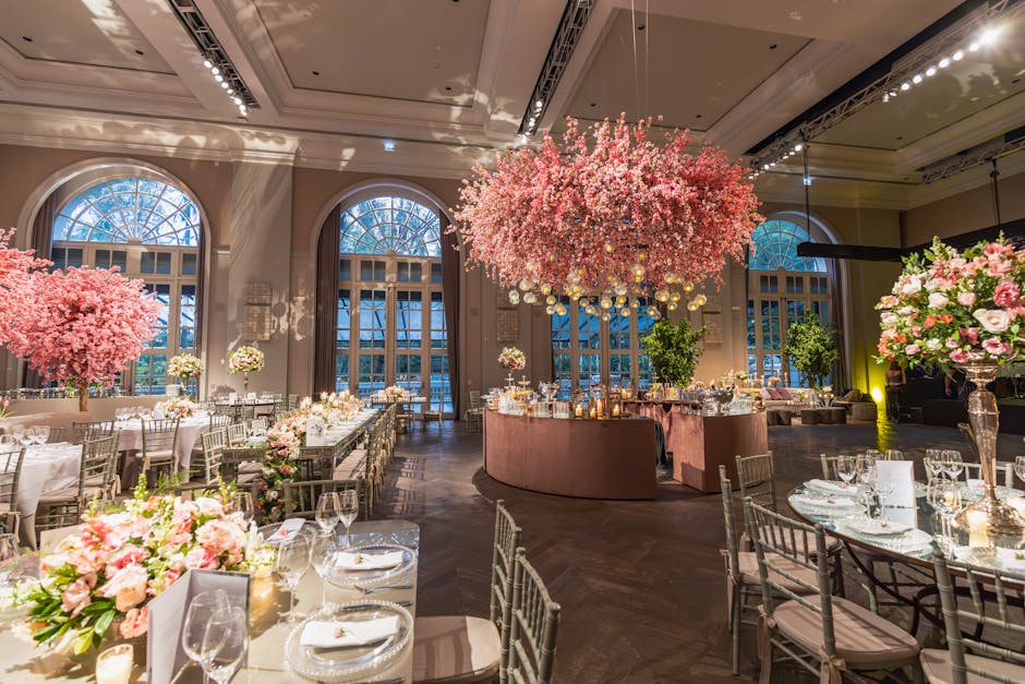 An elegant event space illuminated by soft, natural light from large arched windows, featuring a grand central floral display suspended from the ceiling, composed of densely packed pink cherry blossom
