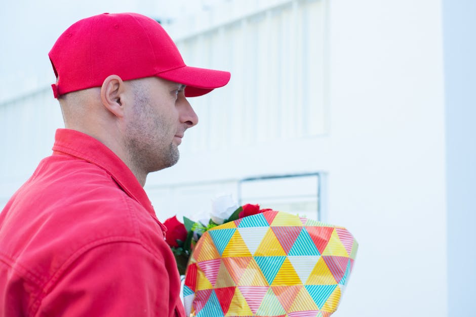 A delivery person wearing a red cap and matching red jacket holding a floral gift wrapped in colorful, geometric-patterned paper with a yellow, blue, and red triangle design, standing outdoors near a