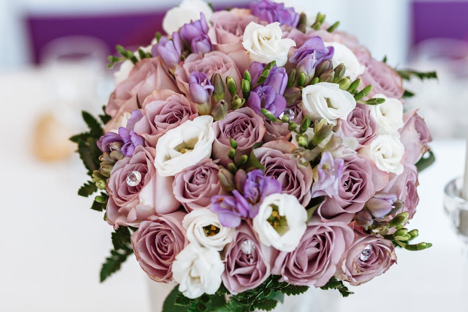 A close-up of a floral arrangement featuring a bouquet of soft pink and lavender roses, interspersed with white lisianthus and purple freesia, accented with small green foliage. The flowers are fresh,