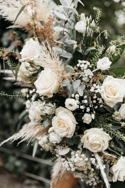 A close-up of a floral arrangement featuring cream-colored roses, small white lisianthus, baby's breath, and textured greenery such as eucalyptus and dried grasses. The flowers are artistically arrang