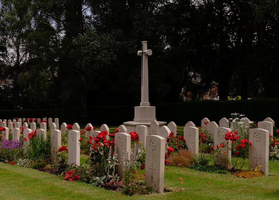 A memorial garden featuring a collection of white gravestones arranged in neat rows, each engraved with symbols and inscriptions. In the center stands a white cross on a pedestal, surrounded by vibran