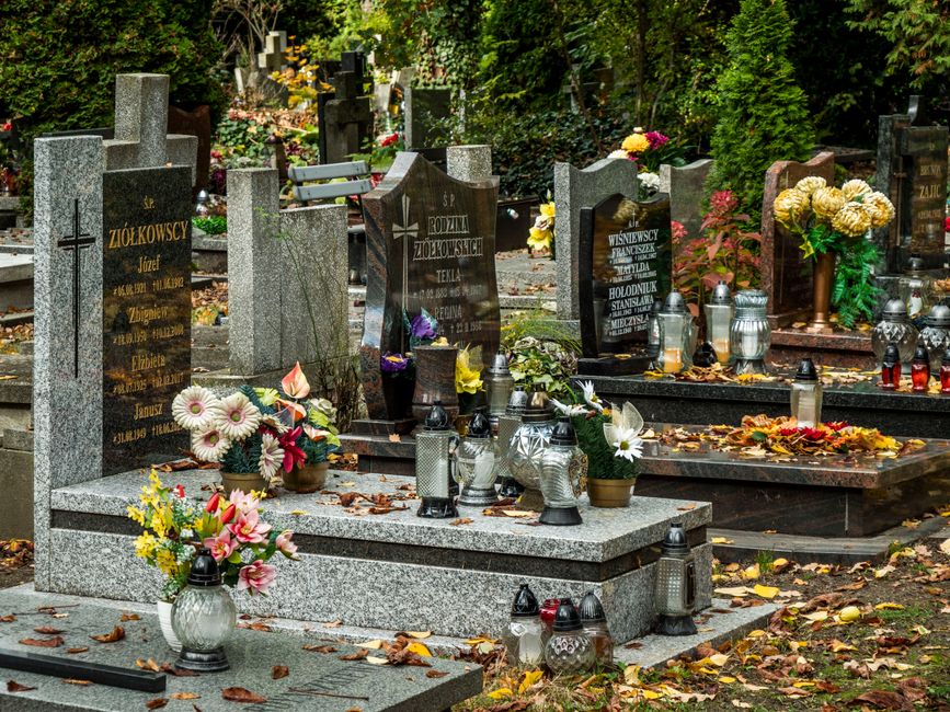A serene cemetery scene featuring several polished granite headstones surrounded by lush green trees and shrubs. In the foreground, a bouquet of fresh flowers, including pink and white daisies, tulips